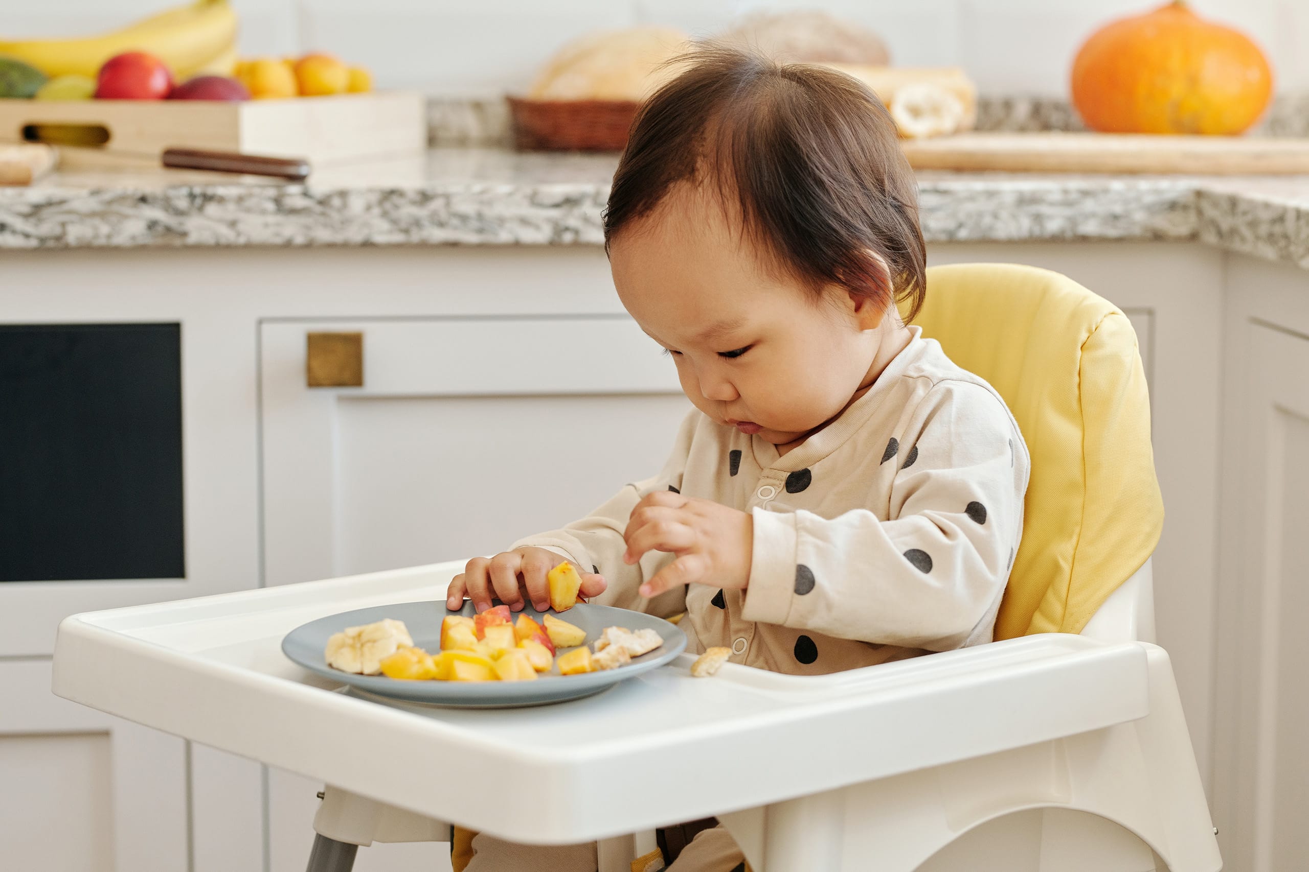 Adorable Asian baby enjoying sliced fruits for breakfast in a baby chair within a homely kitchen setting, showcasing Travel'N Tots high quality mealtime rental equipment