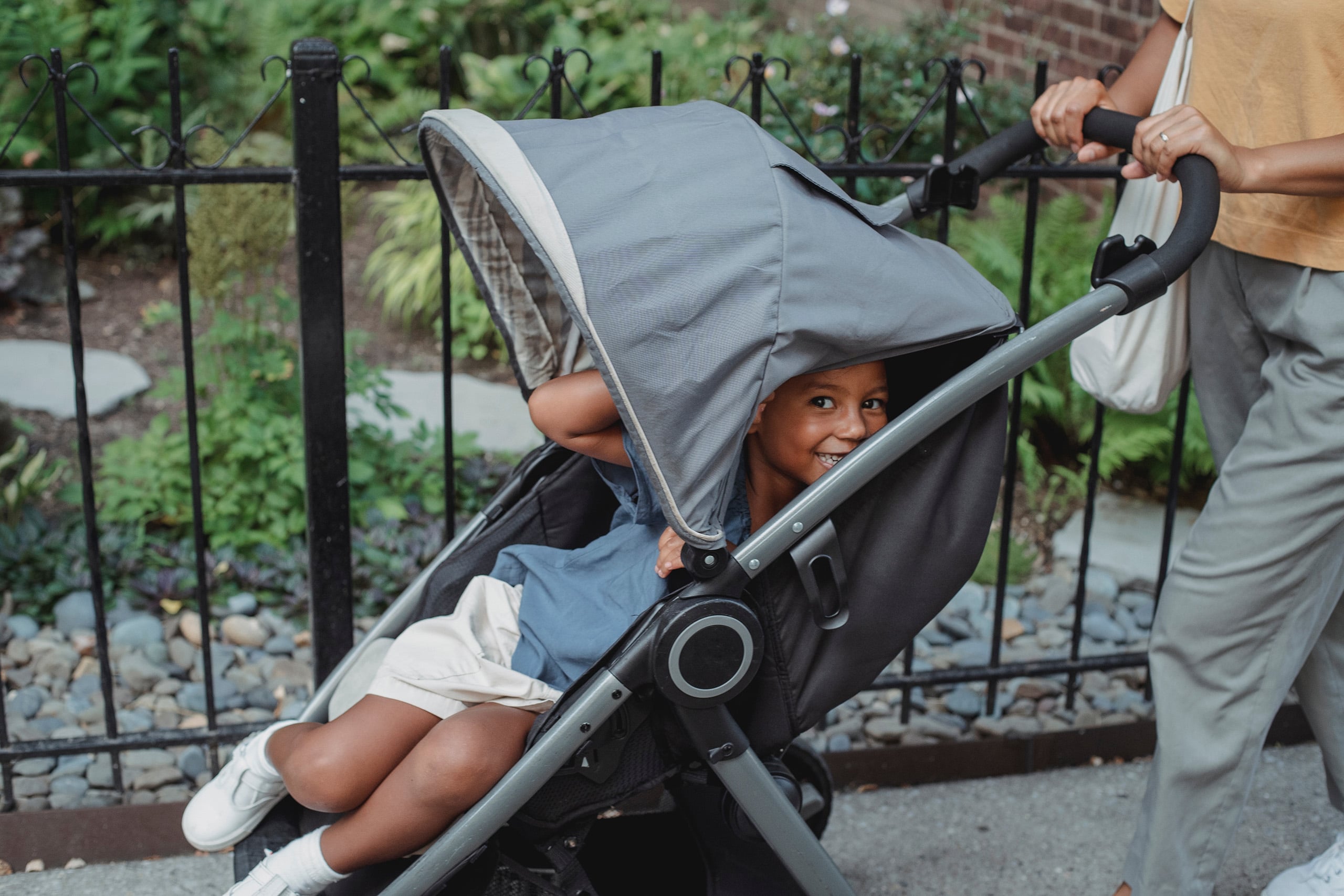 Cheerful colored boy snugly seated in a stroller with a protective hood, handled by his mother on a sidewalk during a cloudy day, representing Travel'N Tots convenient travel time rental solutions