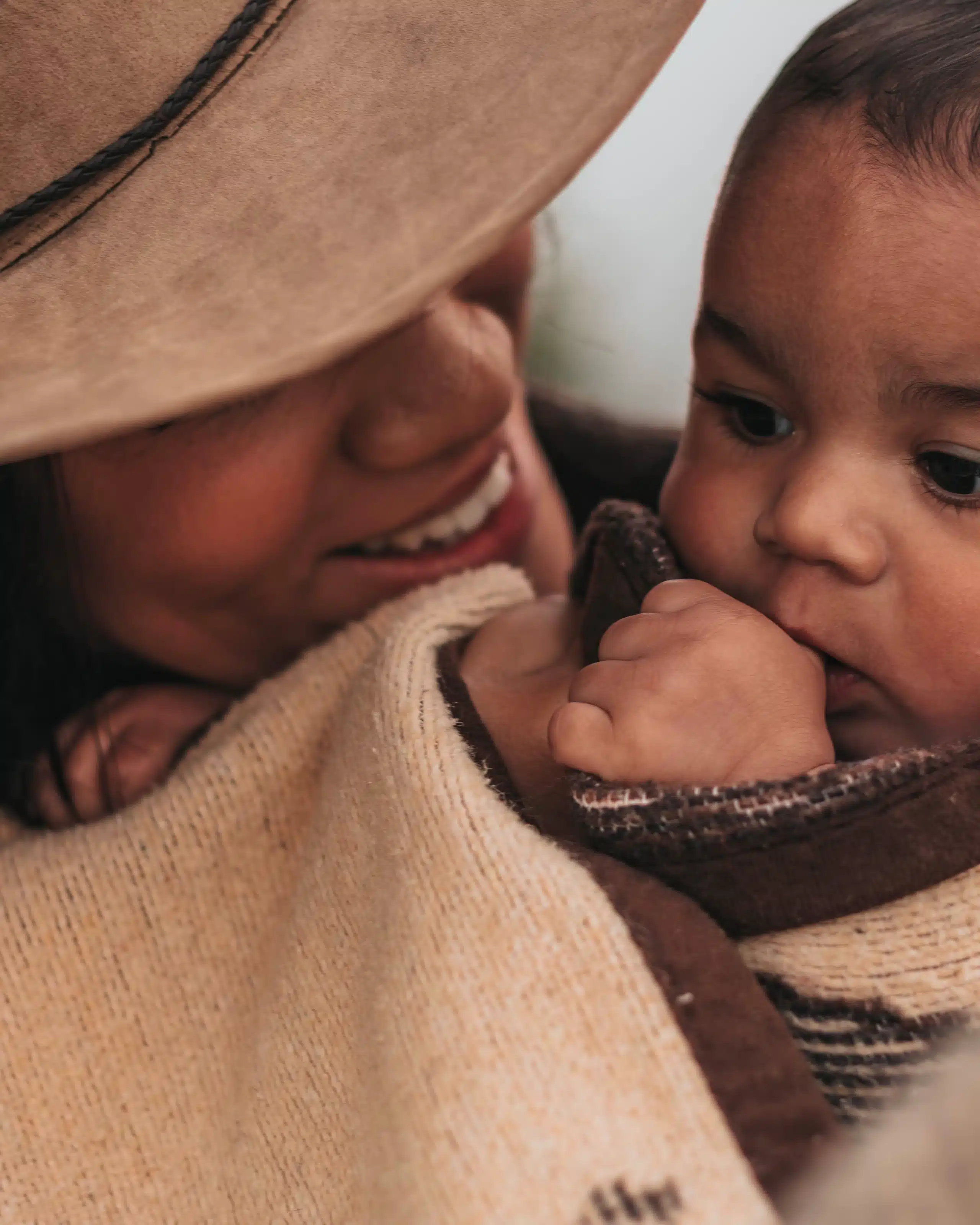 Diverse mother wearing a hat, tenderly covering herself and her baby with a blanket in the morning light, epitomizing motherhood and the nurturing essence of Grapevine Placement Agency