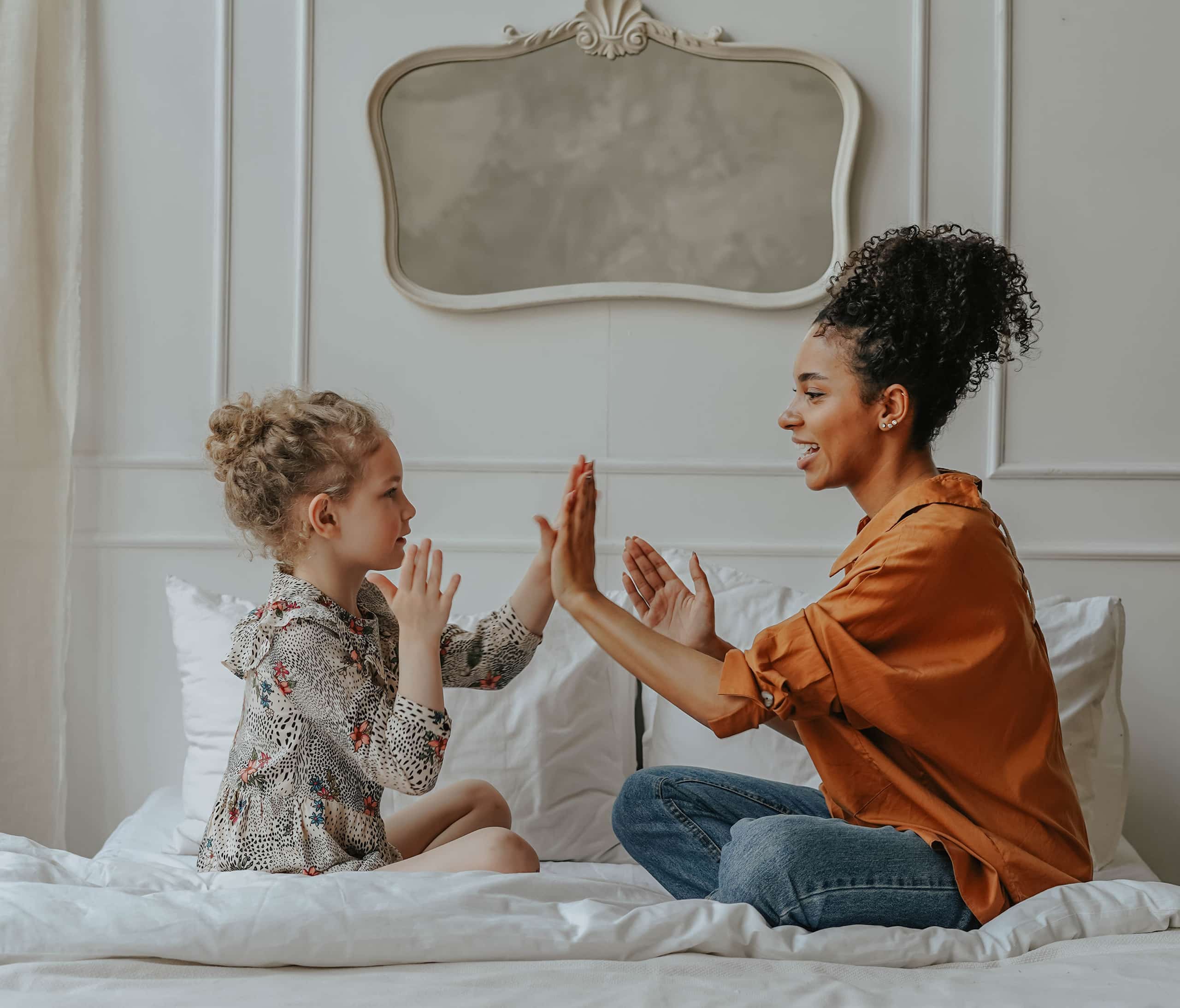 Diverse nanny engaging in hand games with a white young girl on a pristine bed in a luxurious hotel room, embodying Grapevine Placements Agency's specialized hotel guest services