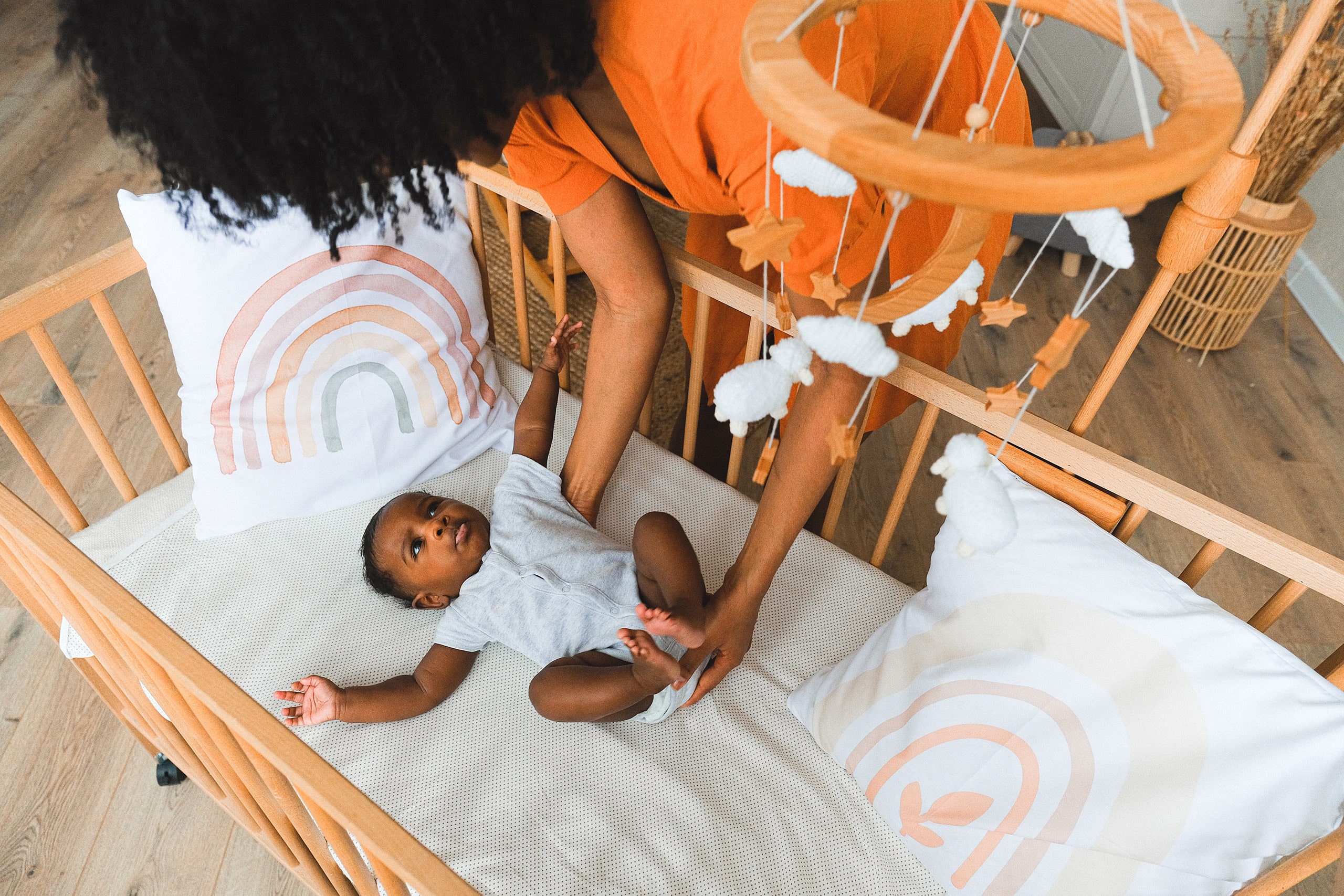 Loving colored mother gently placing her baby in an upscale wooden crib adorned with rainbow cushions and a whimsical mobile, highlighting Travel'N Tots premium bedtime equipment for rent