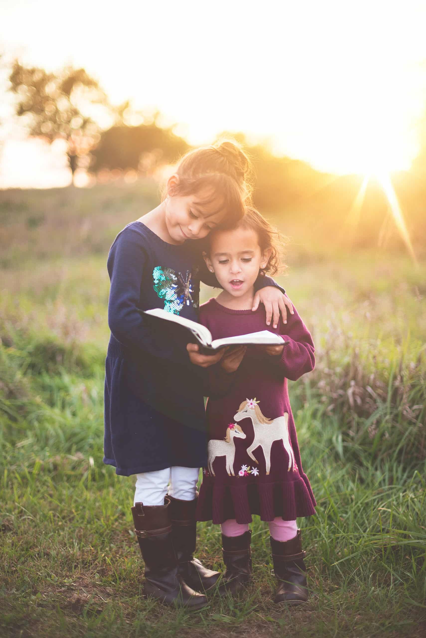 Two young girls engrossed in reading a book together, standing on a lush green lawn.